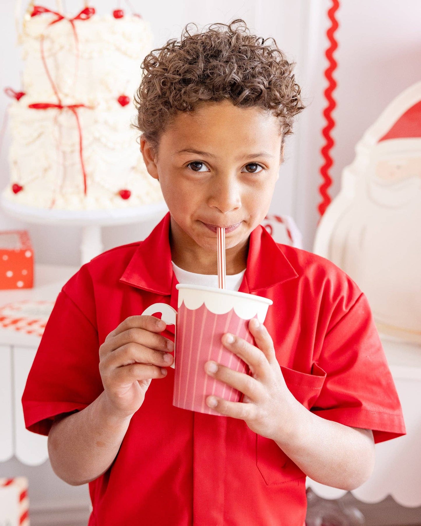 Child in a red shirt drinking from a pink cup with a straw, Christmas-themed background