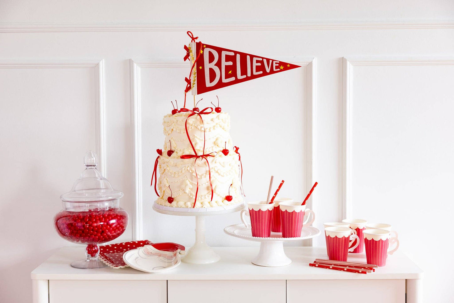 Decorative cake with 'BELIEVE' flag on a white surface with red and pink decorations.