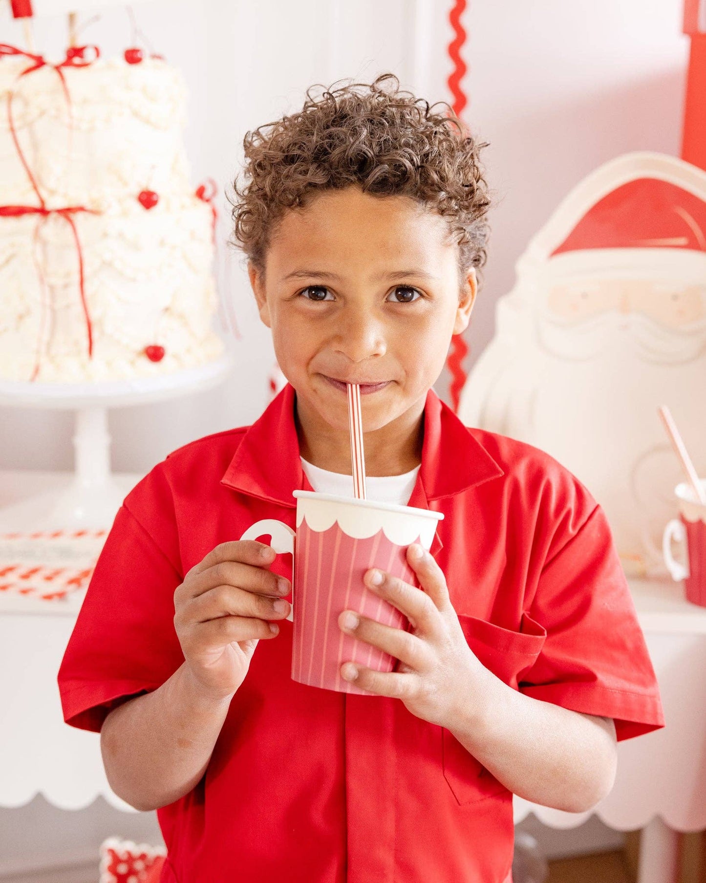 Child in a red shirt holding a Christmas-themed cup with a straw, with a festive background.