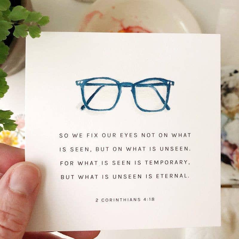 Card with blue eyeglasses illustration and 2 Corinthians 4:18 text held by a hand.